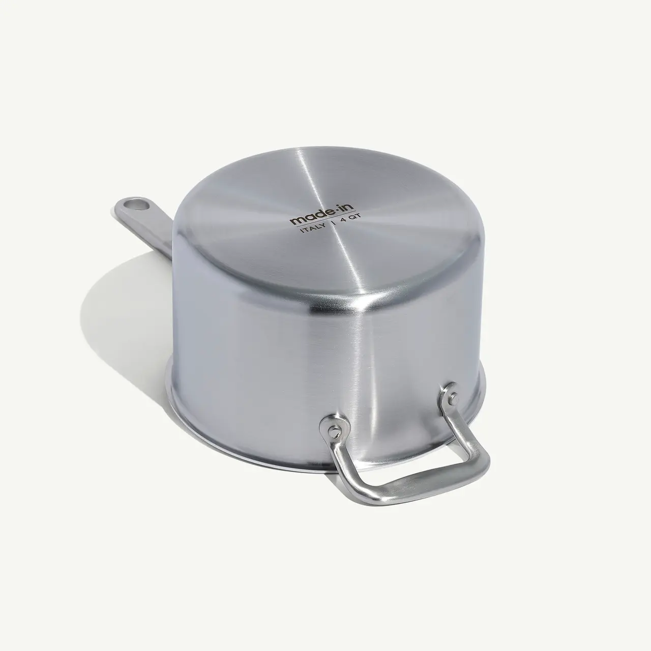 A stainless steel saucepan with a lid and a long handle casts a shadow on a light surface.