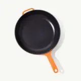 A black non-stick frying pan with an orange handle is shown from above on a pale background.