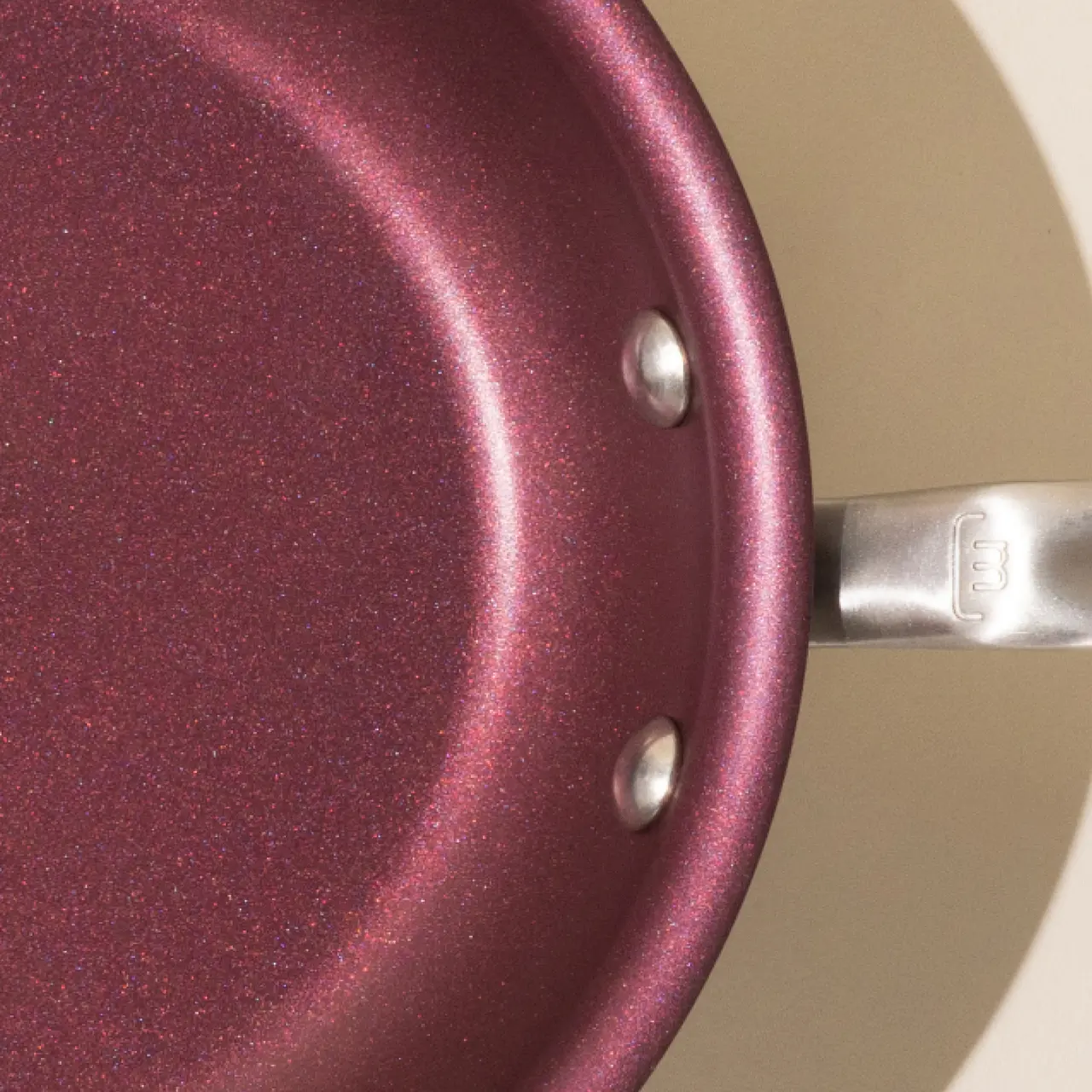 A close-up view of a burgundy non-stick frying pan with a metallic handle attached by two rivets.