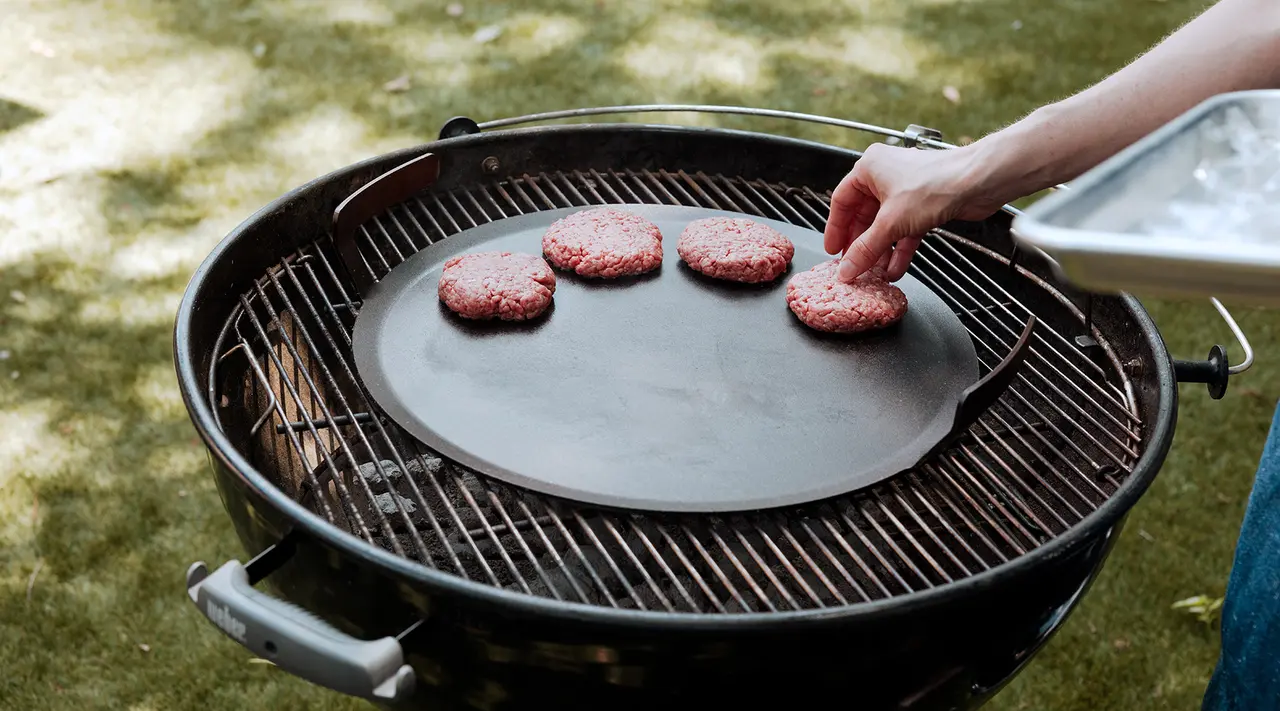 A person places burgers on a circular flat top grill set within a round barbecue over a grassy background.