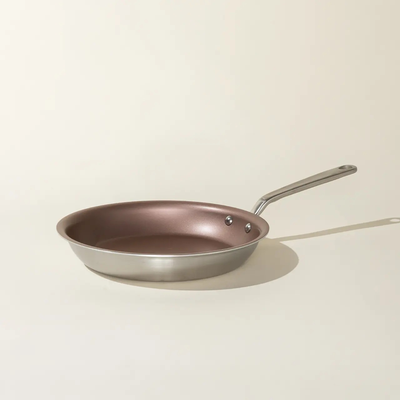 A single stainless steel frying pan with a copper-colored interior and a long handle is positioned against a neutral background.