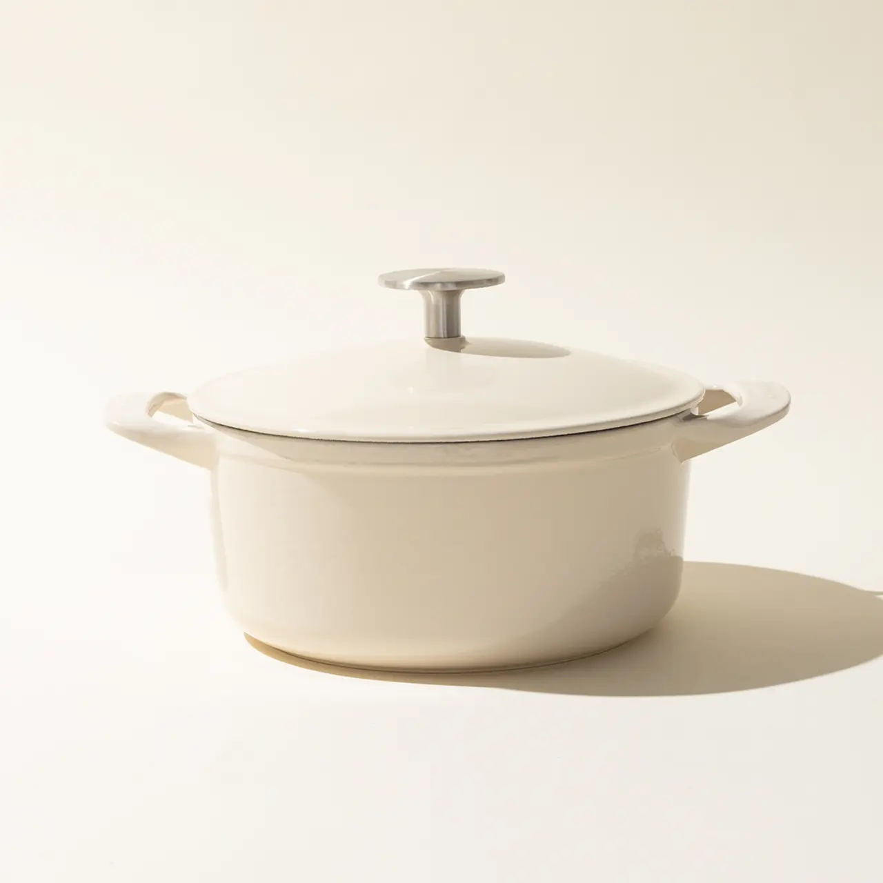 A cream-colored cooking pot with a lid on a light neutral background, casting a soft shadow.