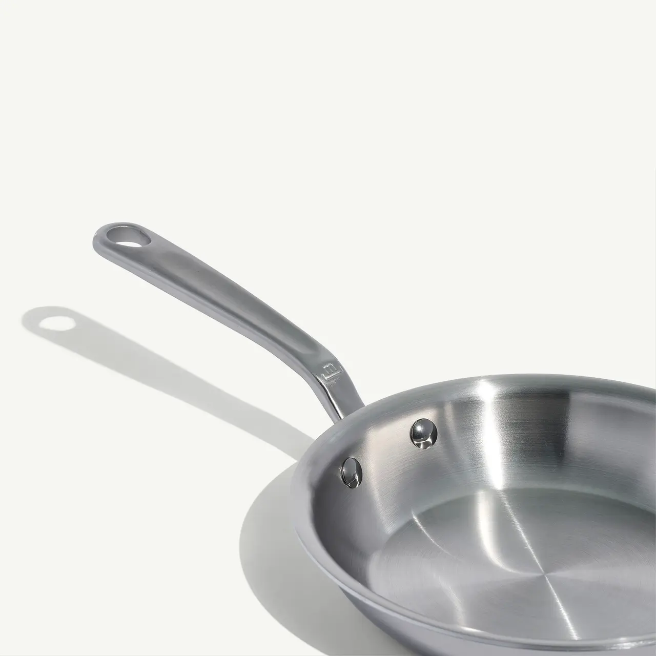 A stainless steel frying pan is positioned on a plain background, casting a soft shadow to the right.