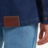 A close-up view of a person's hand on the hem of a denim jacket, featuring a leather label that reads "Wrangler MADE IN."