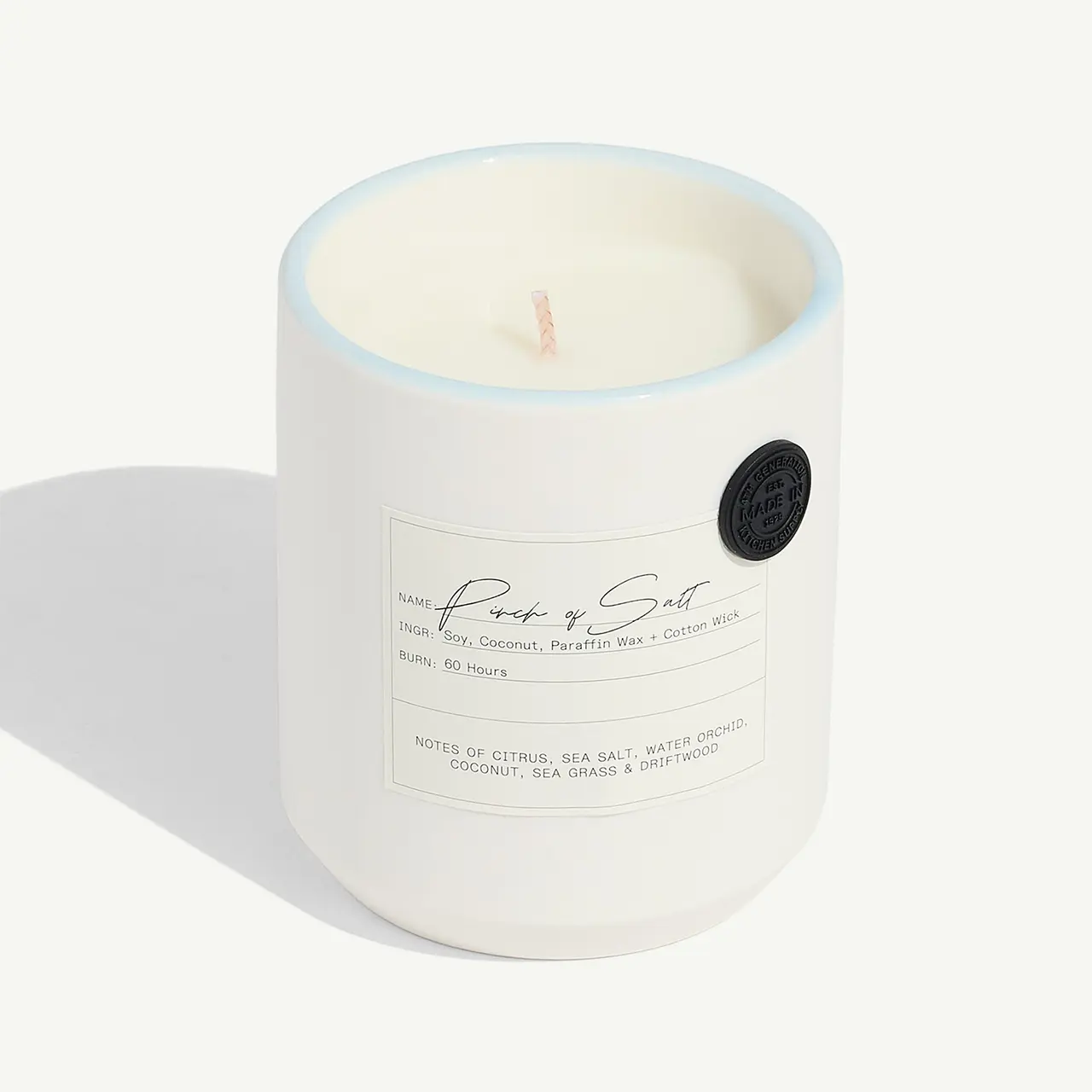 A scented candle with a lit wick is displayed, featuring a label and a logo on its white container.