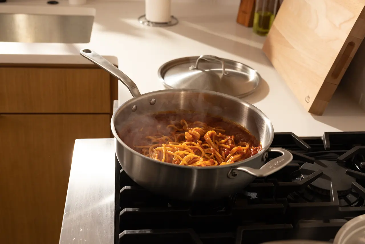 A stainless steel pan with pasta and sauce cooking on a stovetop in a kitchen.