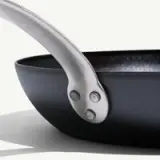 A close-up of a black frying pan shows the detail of its shiny metal handle attached with rivets.