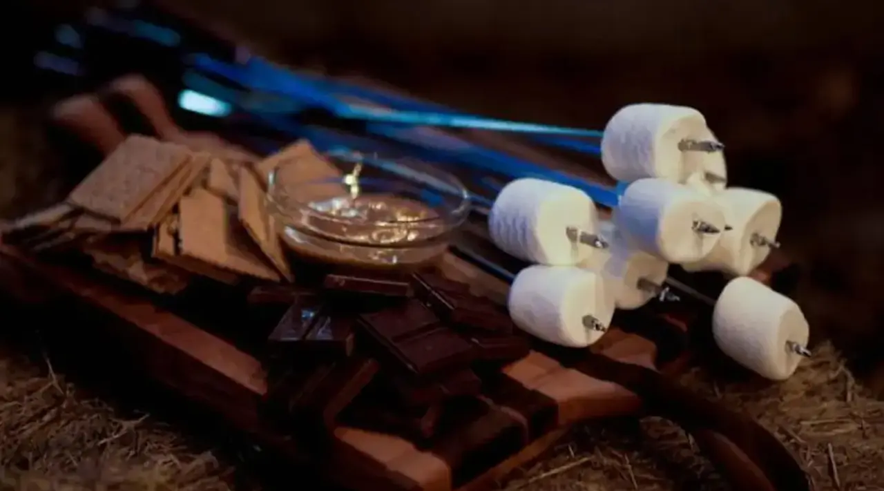 Skewered marshmallows, chocolate bars, and graham crackers are arranged on a wooden surface for making s'mores.