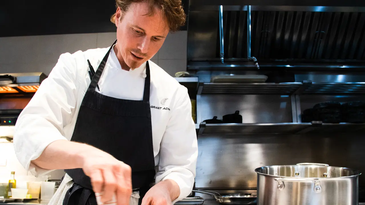 A chef is focused on seasoning a dish while working in a professional kitchen with pots and cooking equipment visible.