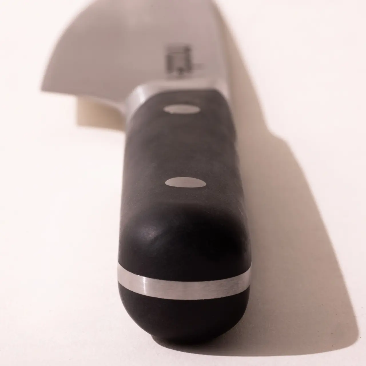 A close-up of a chef's knife with a black handle and three rivets, partially casting a shadow on a light surface.