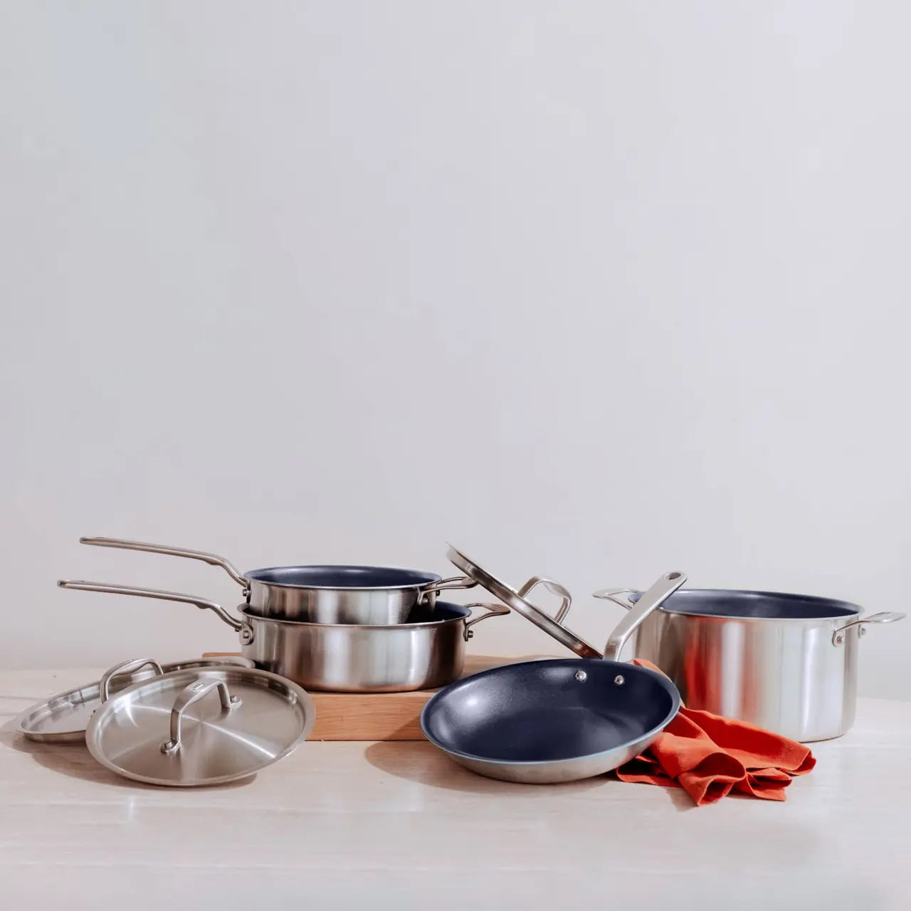 Three stainless steel pots with lids and a frying pan displayed on a wooden surface against a light background, with a red cloth beside them.