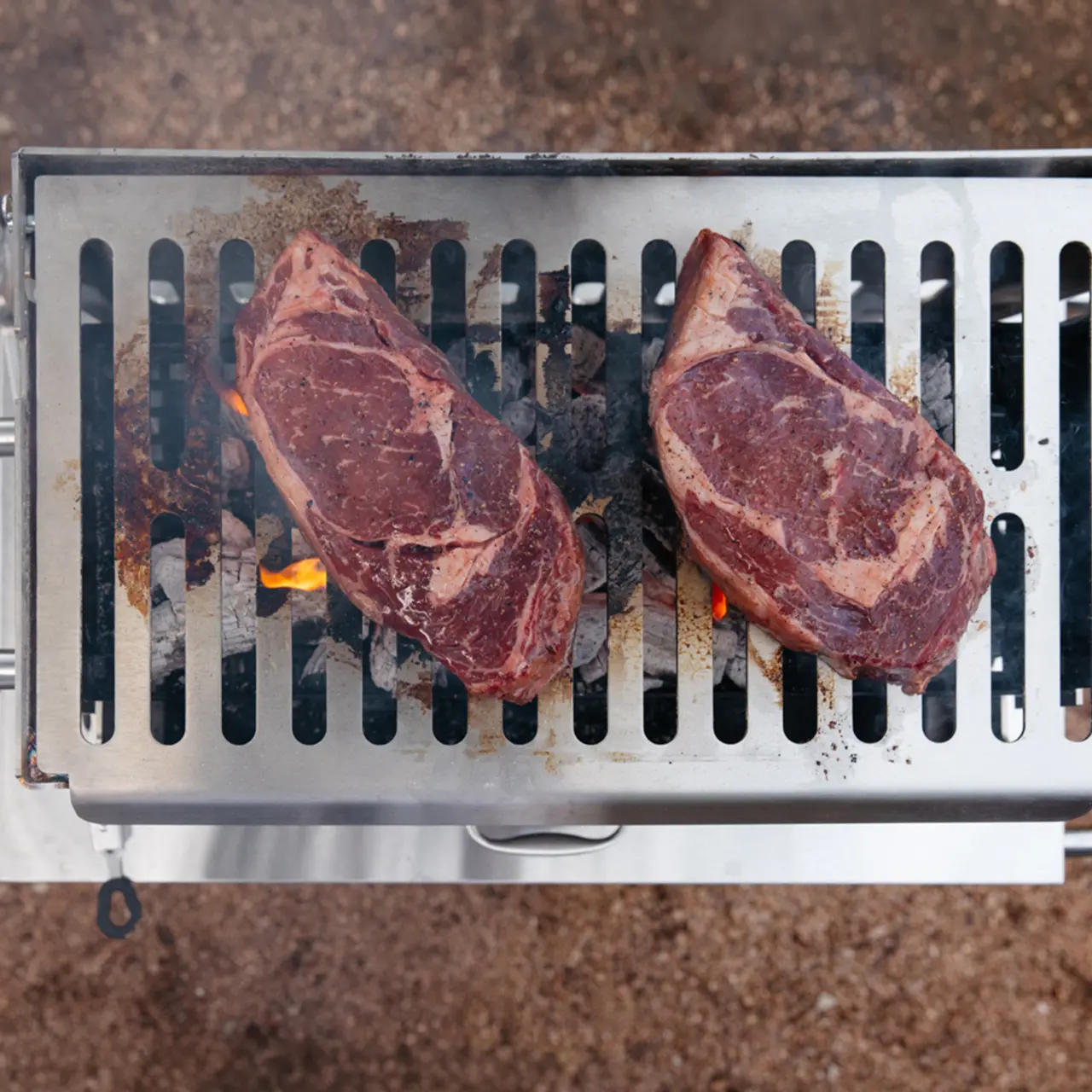 Two thick cuts of steak are sizzling on a grill with visible flames and smoke.