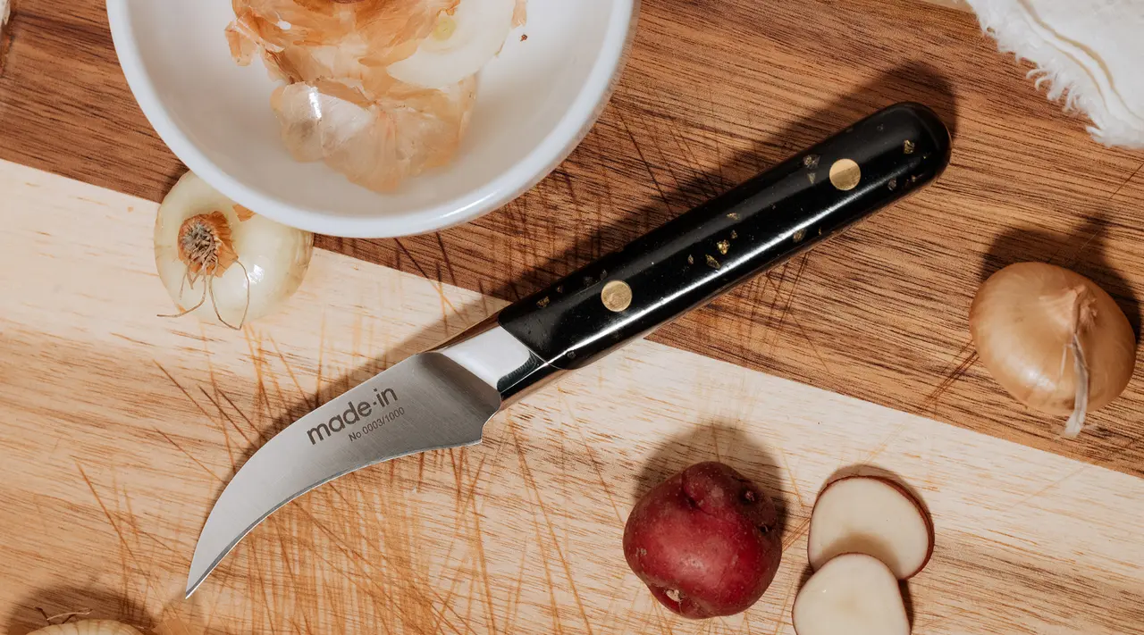 A kitchen knife lies on a wooden cutting board next to sliced potatoes and onion peels.