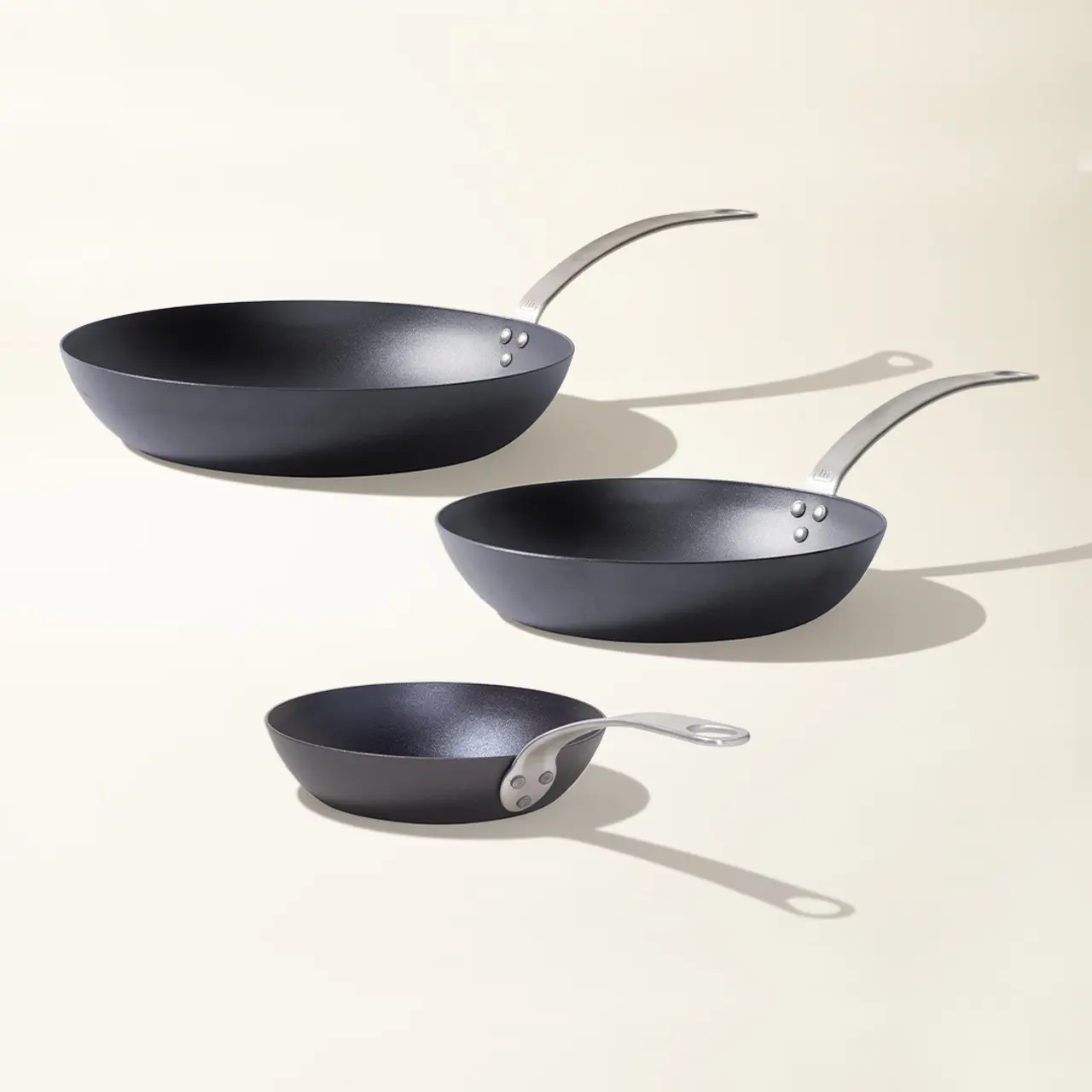 Three different-sized black frying pans are arranged in descending order with their shadows cast on a light background.