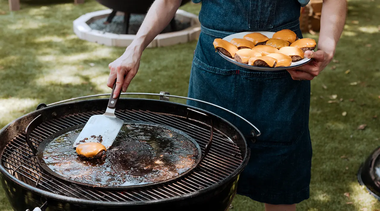 A person in a denim apron flips a burger on a grill while holding a plate of buns nearby, suggesting an outdoor cooking scenario.