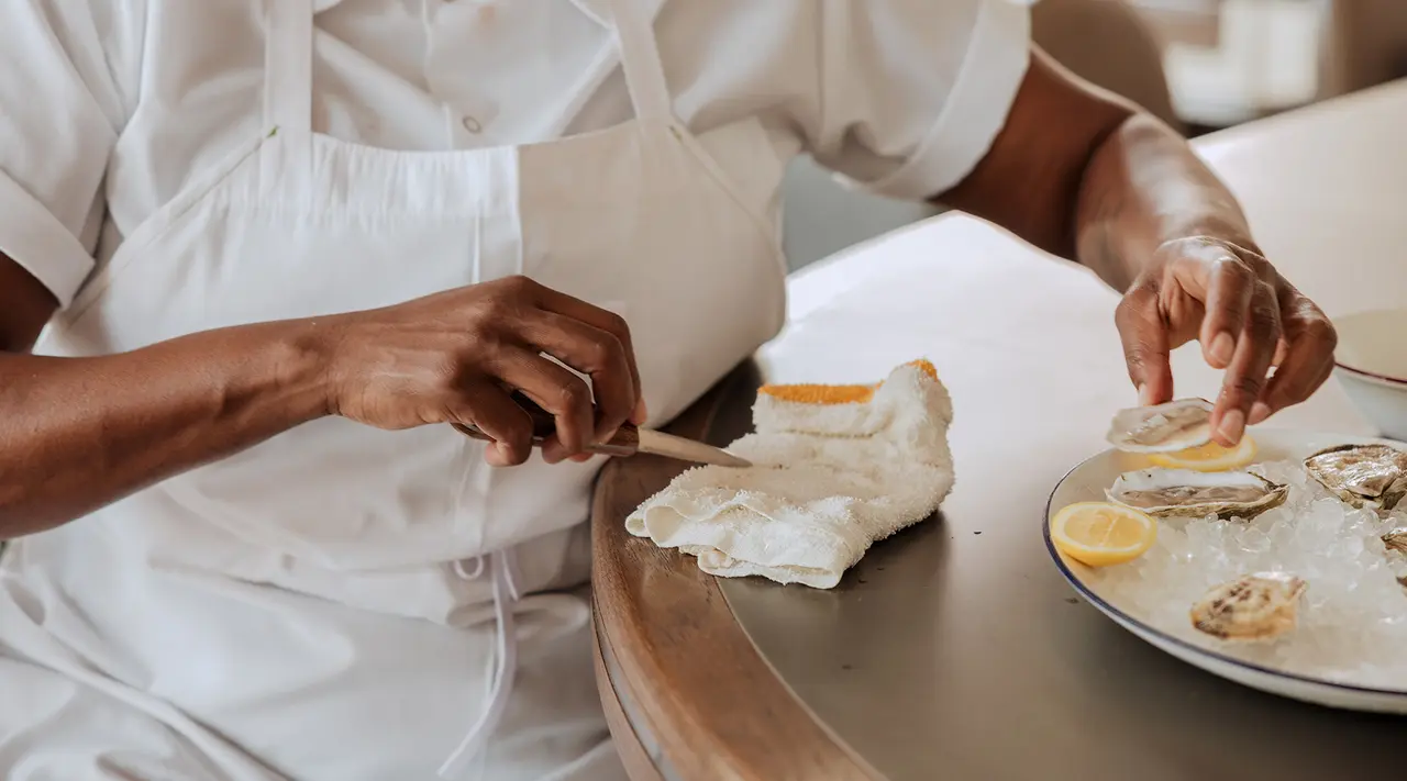 A person in a chef's apron is shucking oysters on a plate accompanied by lemon slices.