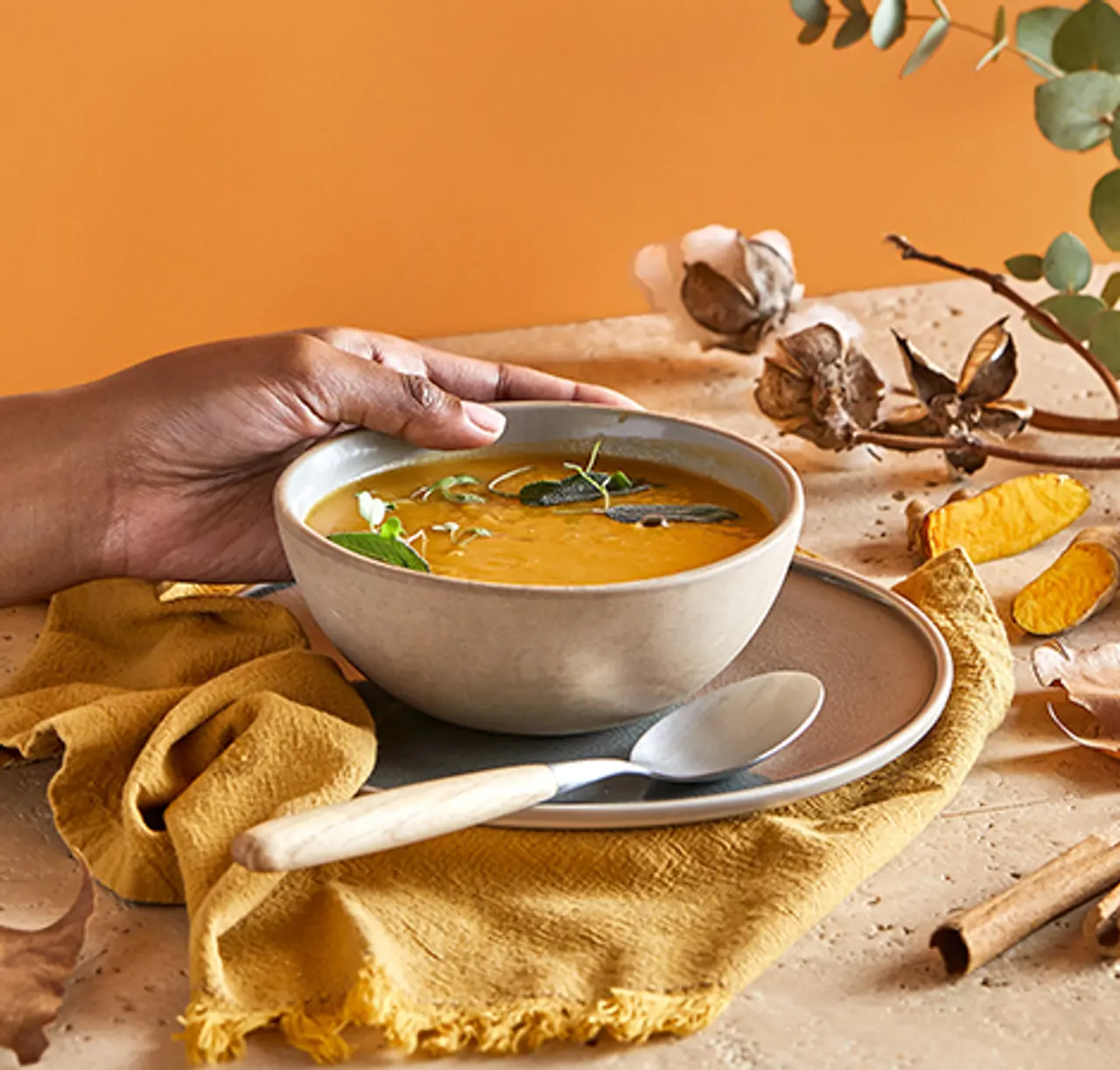 A person's hand is holding a bowl of creamy soup garnished with herbs on a table with autumnal decor.