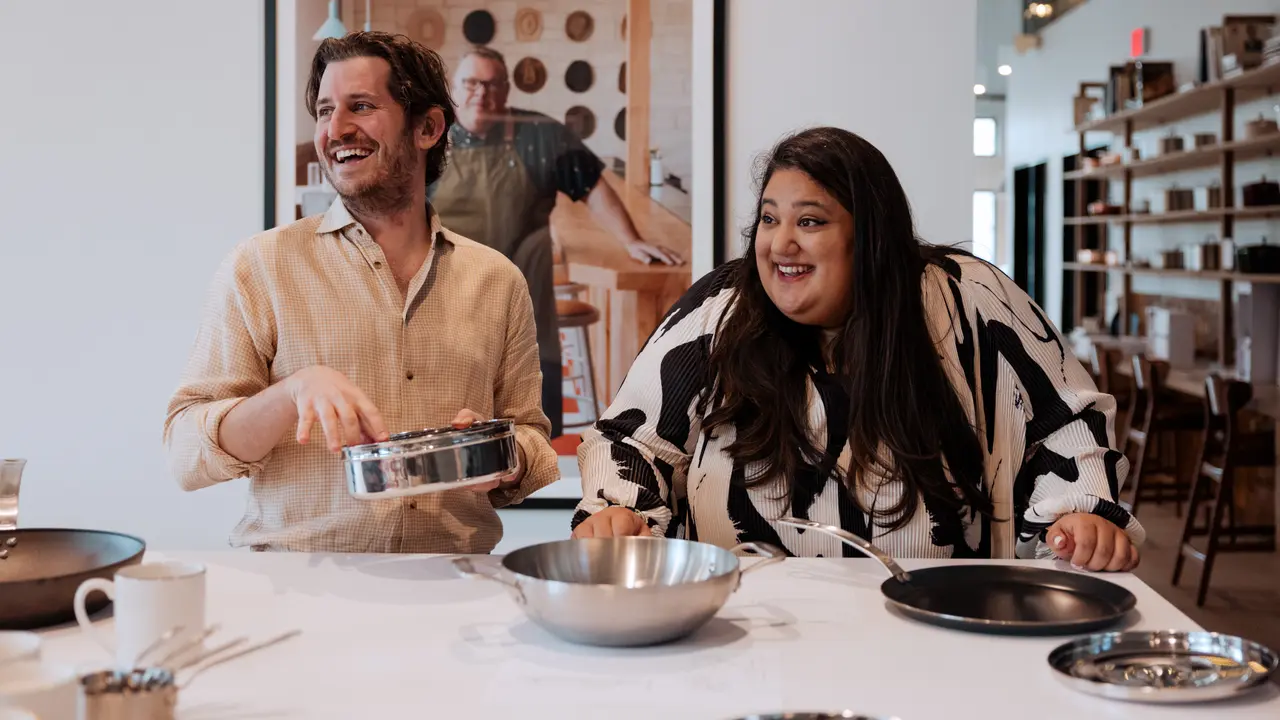 Two people are smiling and engaging in a cooking activity at a table covered with kitchen utensils.