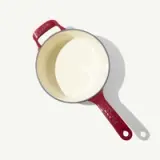 A red cast iron pot with a cream-colored interior and two handles sits against a light background, casting a shadow.