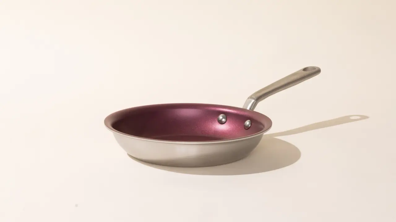 A silver-handled frying pan with a red interior rests on a pale surface casting a slight shadow to the right.