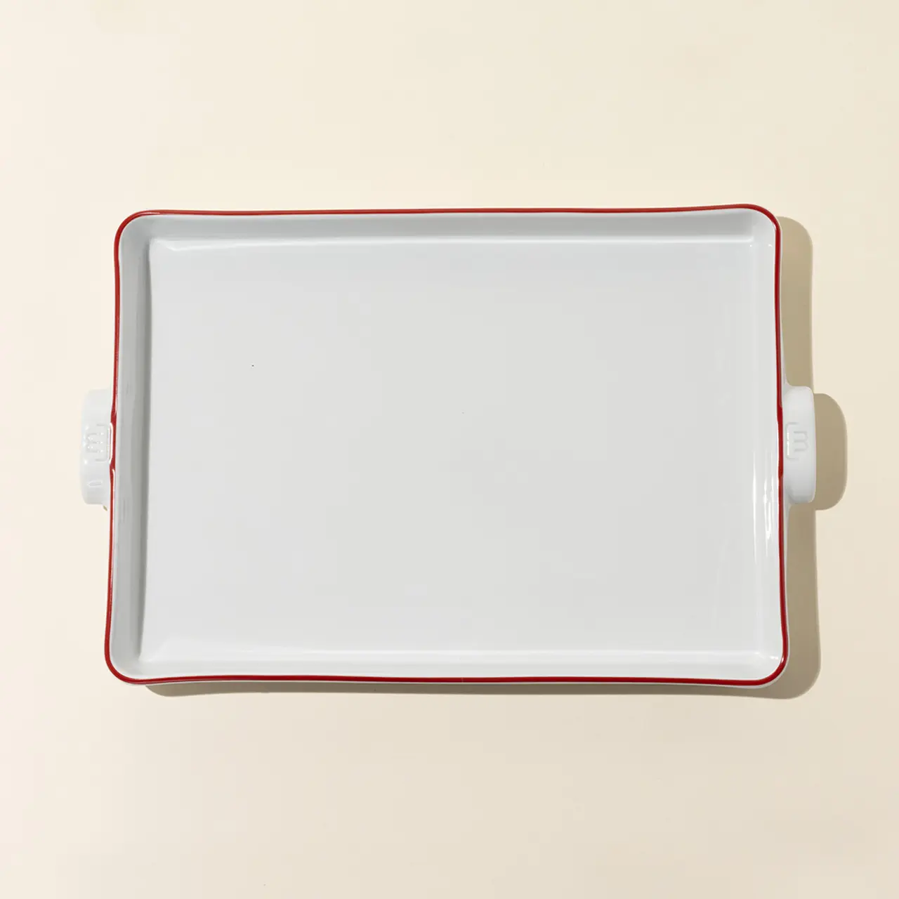 A white square ceramic baking dish with red trim sits on a beige surface.
