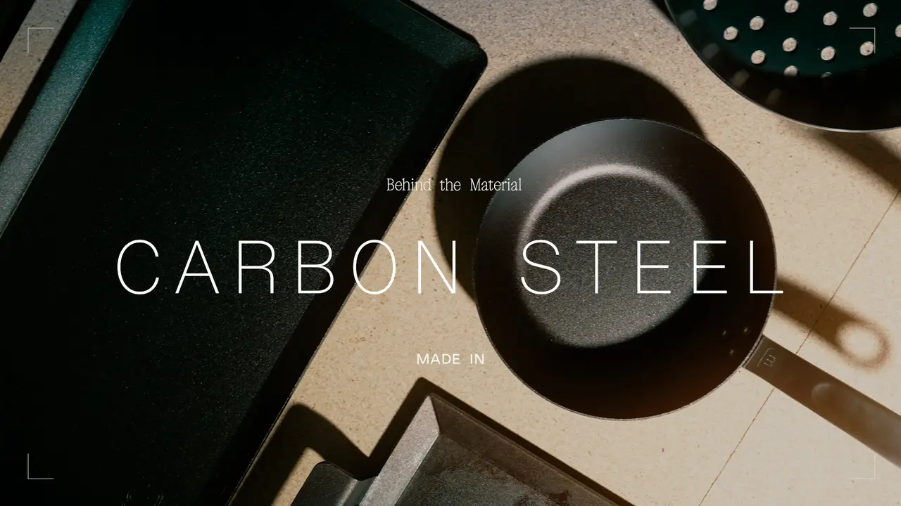 A carbon steel pan and kitchen utensils are set against a textured background, accompanied by text reading "Behind the Material" and "CARBON STEEL MADE IN."