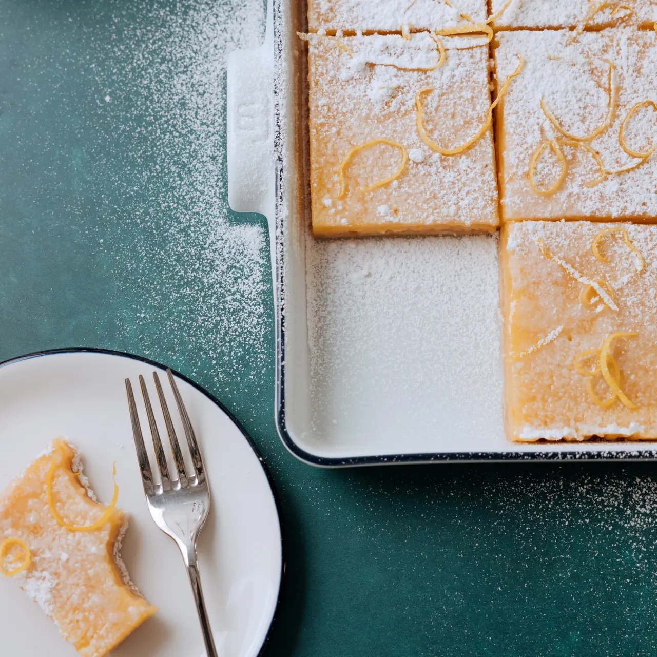 A dusting of powdered sugar tops lemon bars in a baking dish next to a plate with a single bar and a fork on a teal surface.