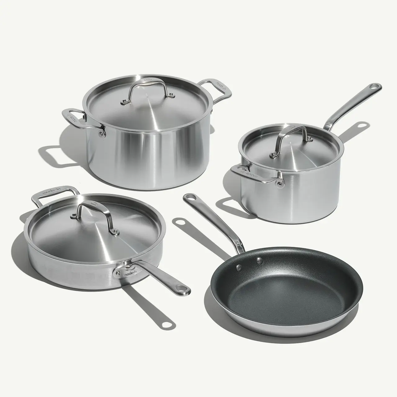 A set of stainless steel cookware with glass lids and one non-stick frying pan on a light background.