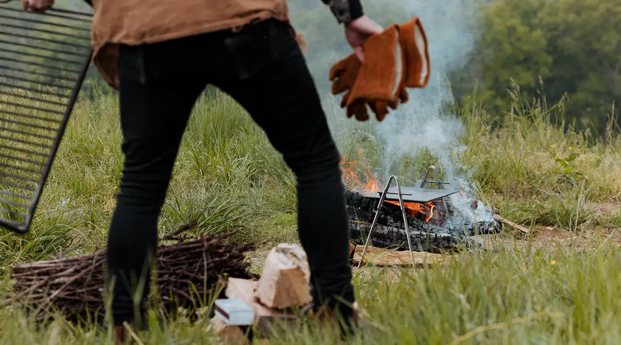 A person in outdoor clothing is standing near a small campfire with smoke rising, holding gloves and preparing to cook or warm up with a metal grill to one side and chopped wood in the foreground.