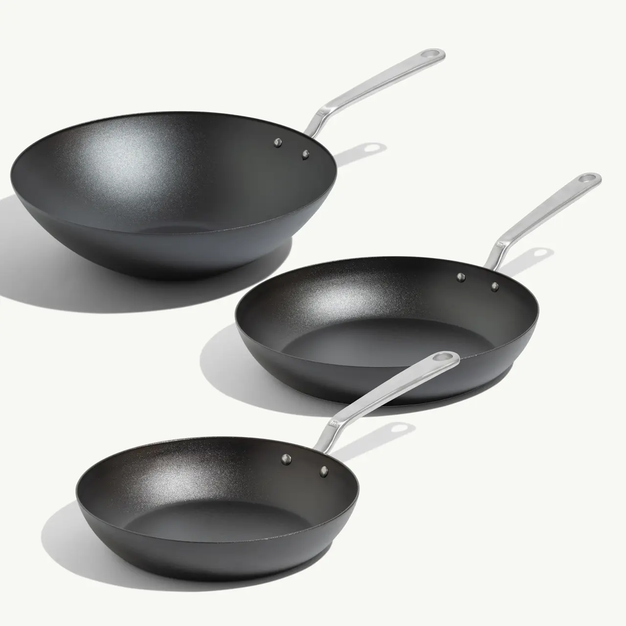 Three black cookware items are displayed, including a wok and two frying pans of varying sizes, all featuring shiny metal handles.