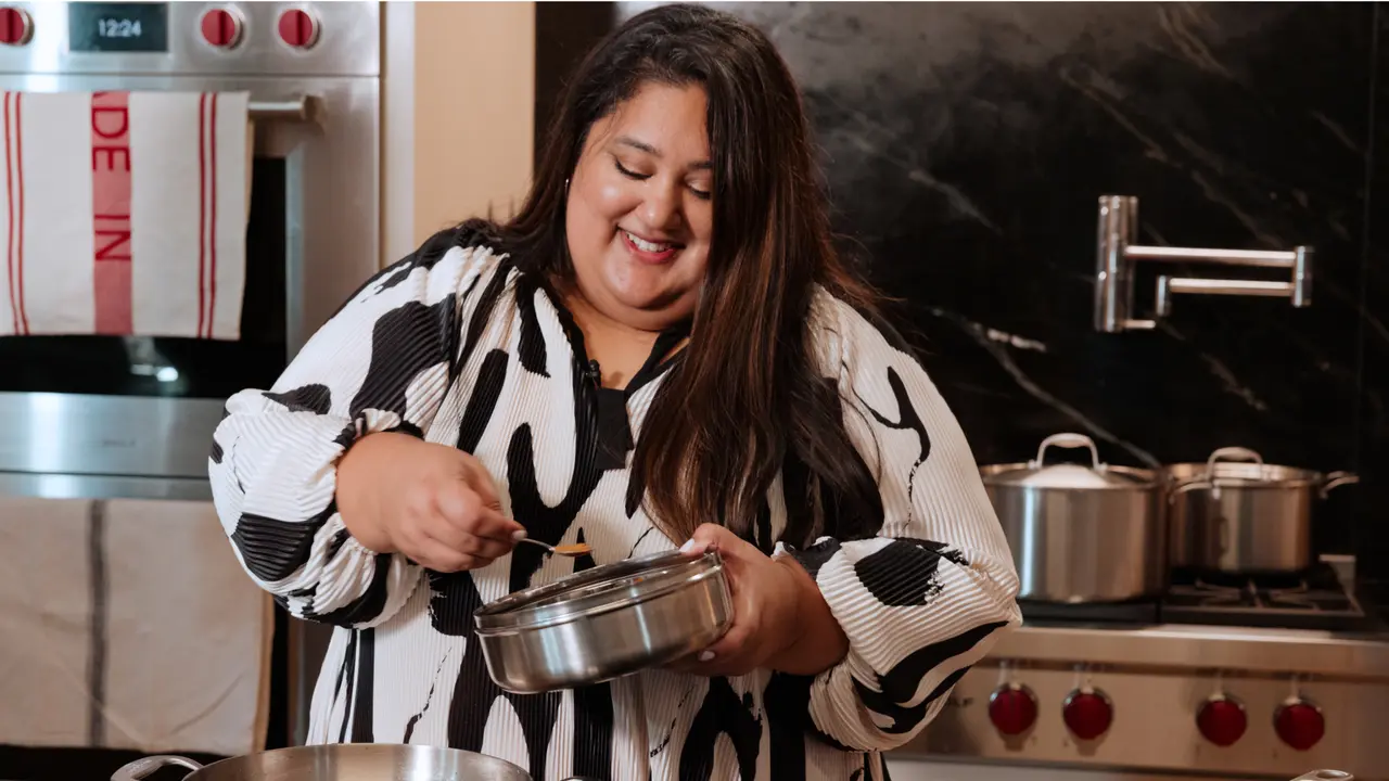 A smiling woman in a patterned top is stirring a pot in a kitchen setting.