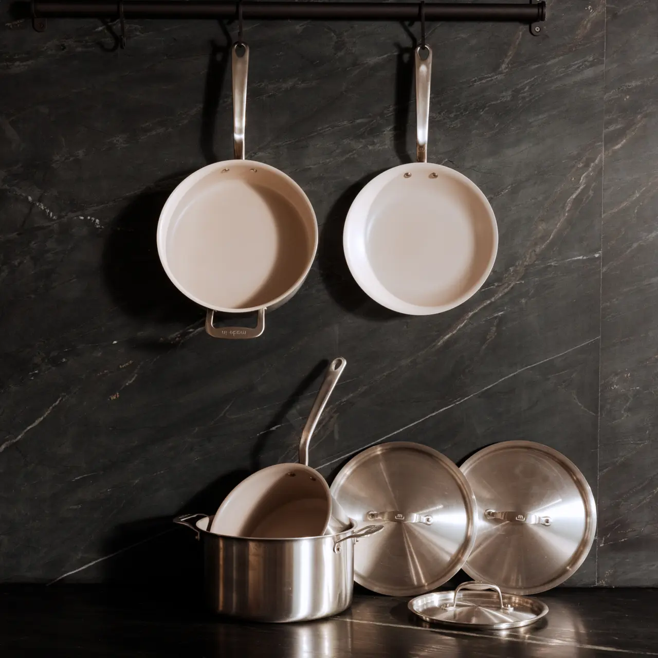A collection of clean cooking pots and pans neatly arranged, with two hanging against a dark tiled wall and three stacked on a countertop.