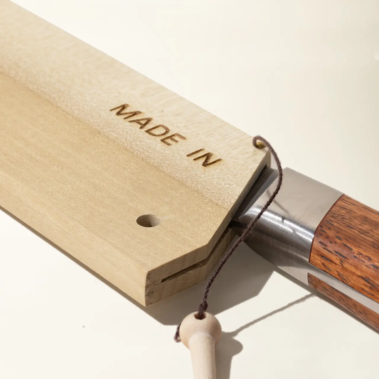 A close-up of a knife with "MADE IN" stamped on light wood, highlighting the blade and wooden handle with a cord through a hole at the end of the handle.