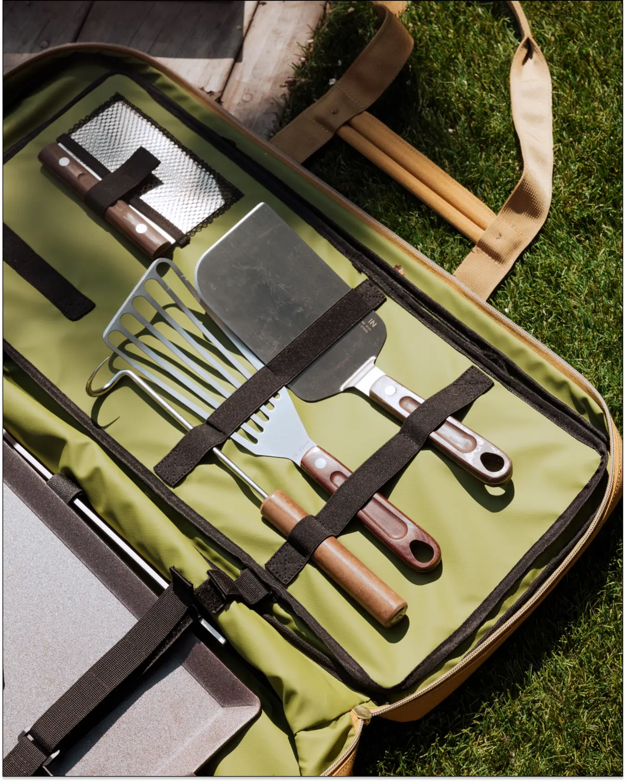 A set of barbecue tools consisting of a spatula, tongs, fork, and two other utensils is neatly organized in a portable, open green case on a grassy surface.