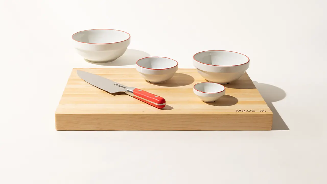 A neatly arranged wooden cutting board displays a kitchen knife and four white bowls with red interiors, casting a soft shadow under bright lighting.