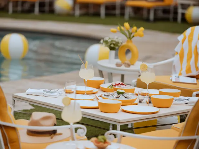 A poolside dining setup featuring a neatly arranged table with yellow and white themed decorations and refreshments under a soft, natural light.