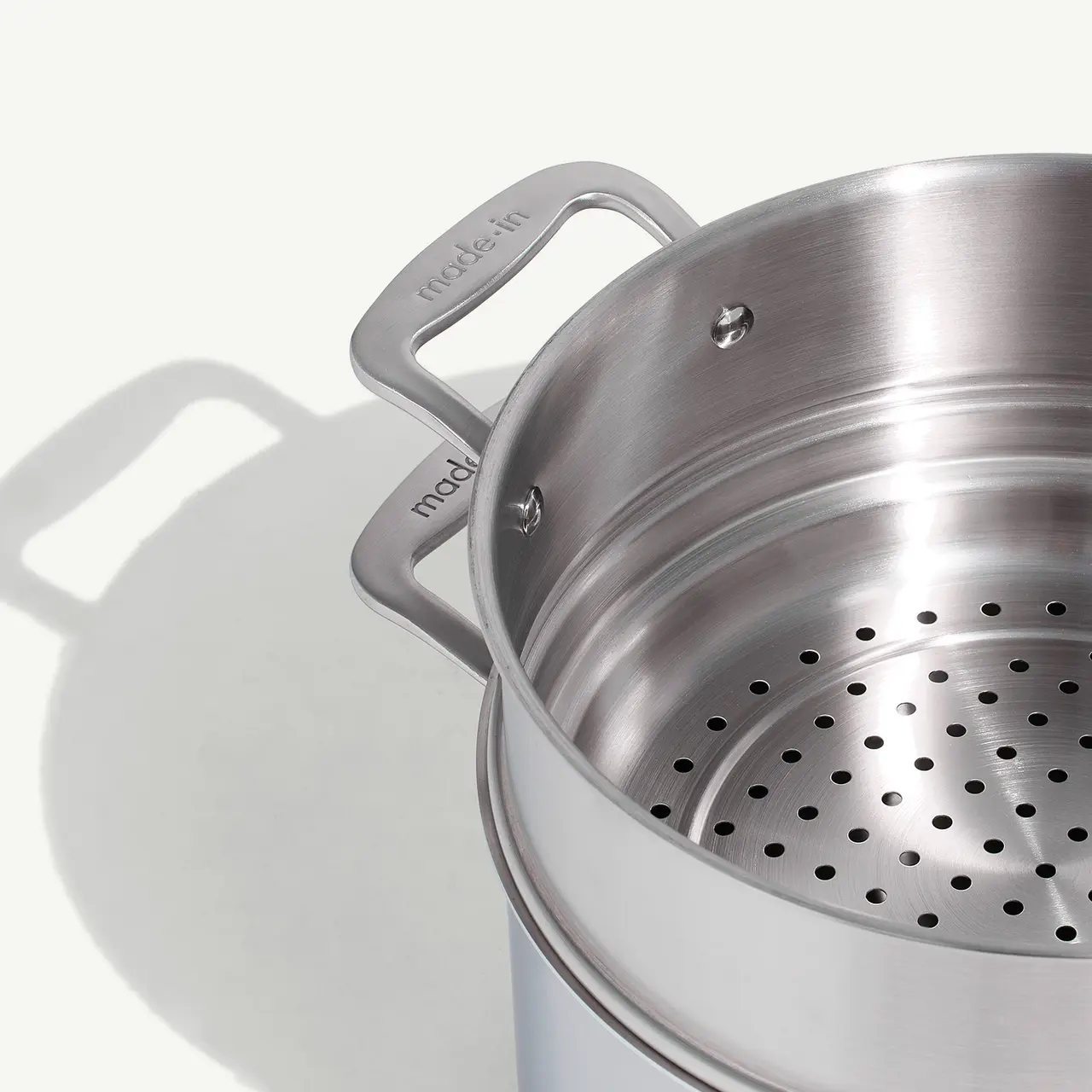A stainless steel pot with a steamer insert is casting a shadow on a white surface.