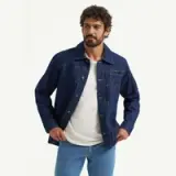 A man with curly hair wears a dark denim jacket over a white t-shirt, posing casually.