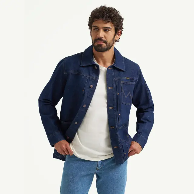 A man with curly hair wears a dark denim jacket over a white t-shirt, posing casually.