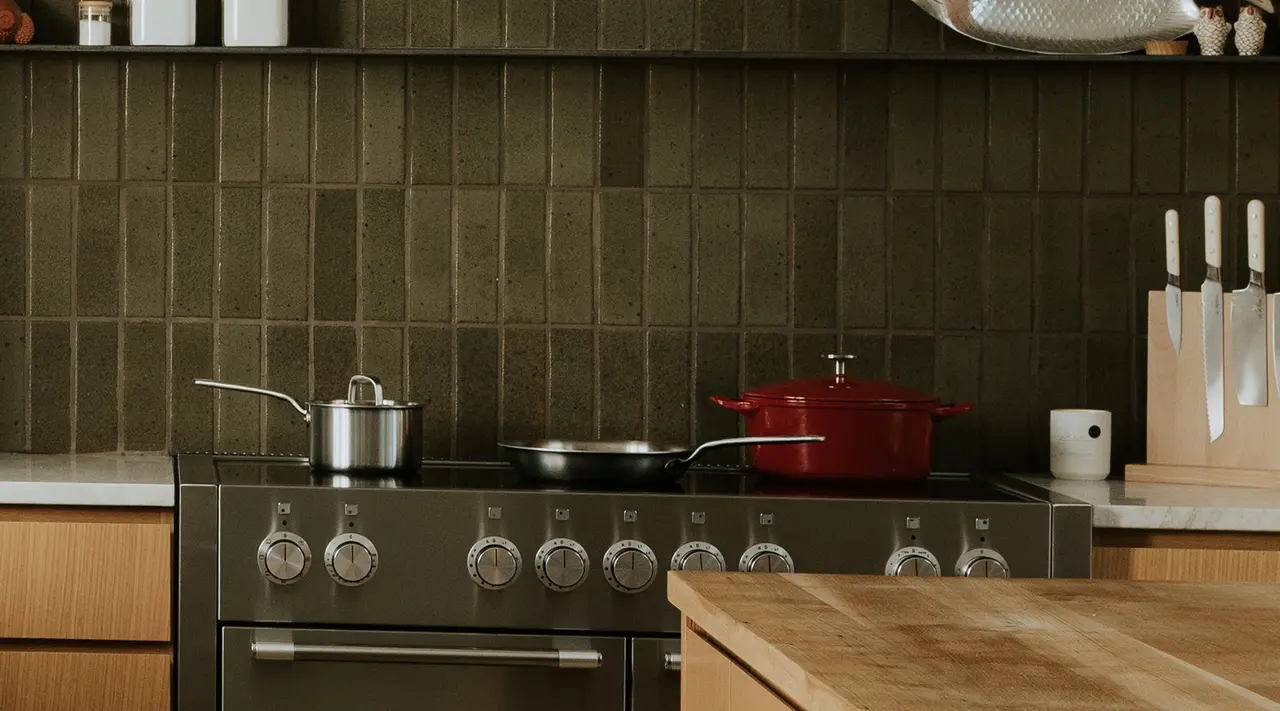 A modern kitchen counter displaying a variety of cookware and a knife set, with a dark tiled backsplash.