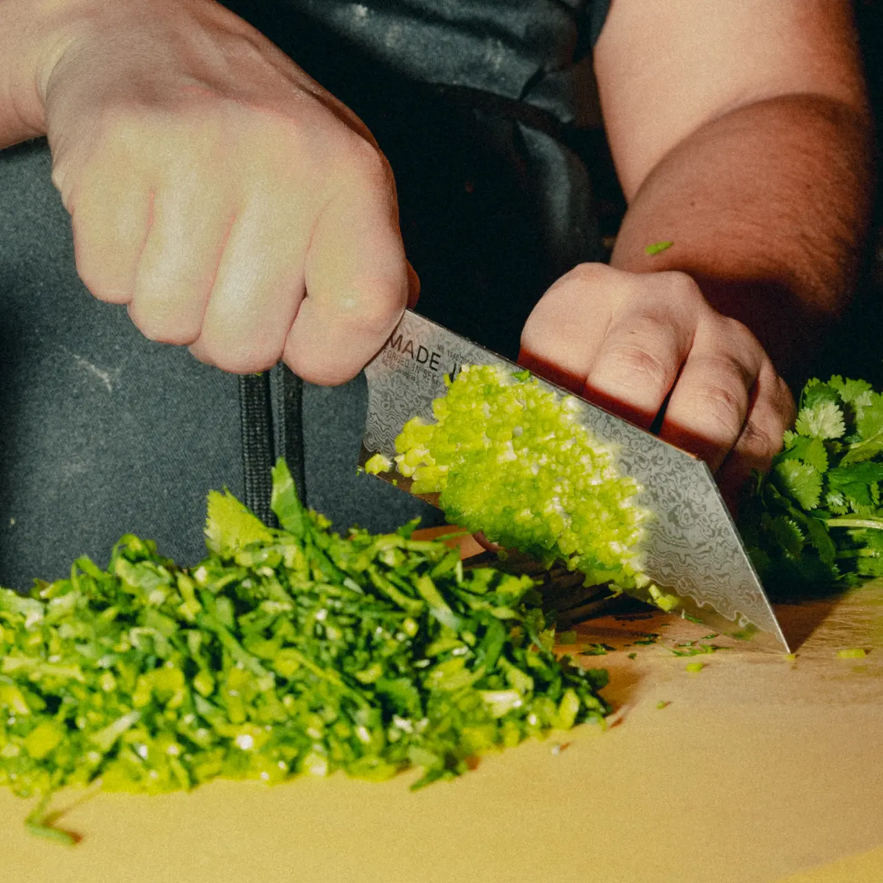 A hand is skillfully chopping fresh green herbs on a wooden cutting board, surrounded by neatly piled chopped cilantro.