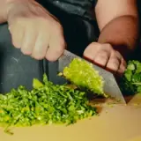 A hand is skillfully chopping fresh green herbs on a wooden cutting board, surrounded by neatly piled chopped cilantro.