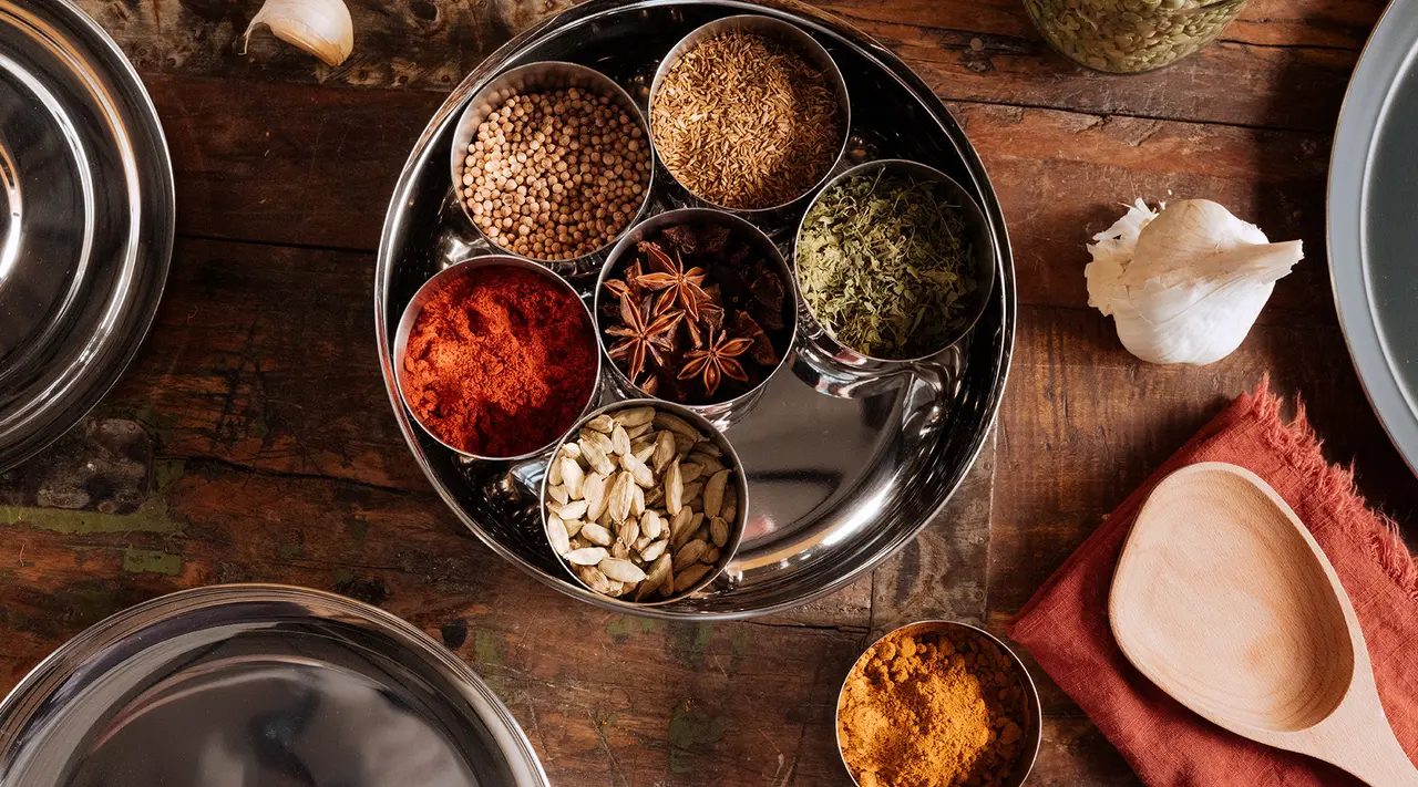 Various spices and herbs are neatly arranged in a stainless steel tray on a wooden table, showcasing a rich palette of colors and textures.