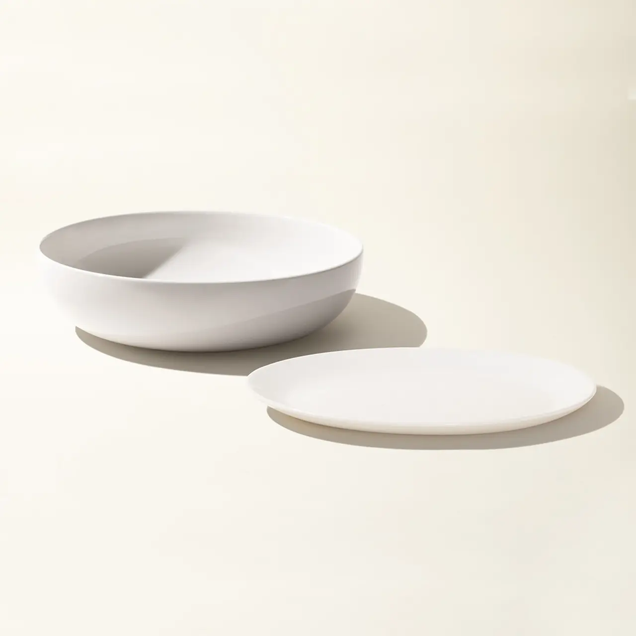 A white bowl and plate are placed on a light-colored surface with a shadow casting to the side.