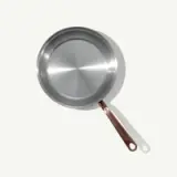 A stainless steel frying pan with a copper handle is positioned against a light background, casting a subtle shadow.