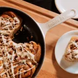 Cinnamon rolls drizzled with icing and topped with nuts are served in a skillet alongside a plated roll.