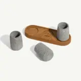 A wooden tray with indentations holds two speckled stone cups, with a third cup lying beside it on a light surface.