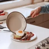 A hand is pouring a fried egg from a skillet onto a slice of toast topped with avocado, alongside crispy bacon on a white plate.