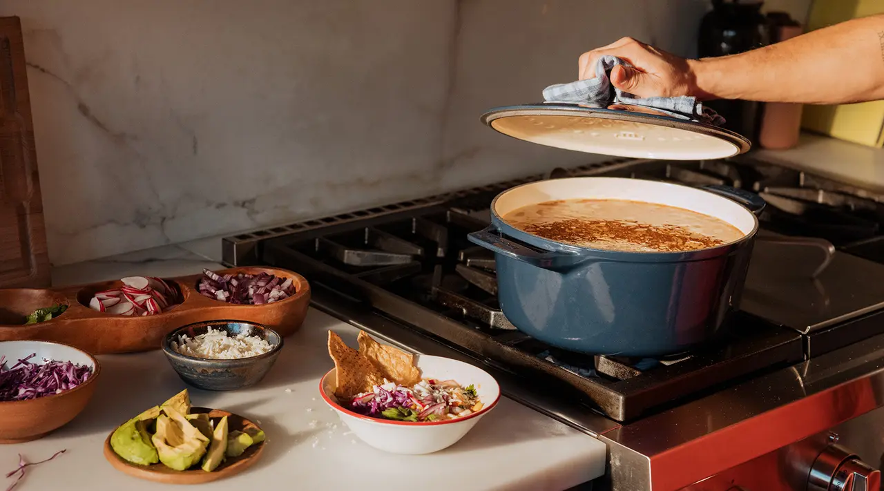 A person lifts the lid off a large pot on a stove surrounded by various ingredients and bowls of food.