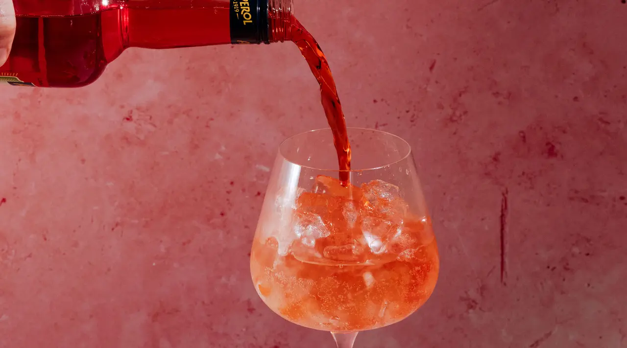 Pouring a refreshing red beverage into a glass filled with ice against a rosy background.